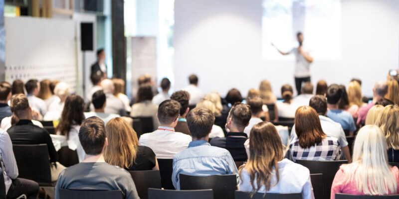 Business speaker giving a talk at business conference event. Speaker giving a talk in conference hall at business event. Audience at the conference hall. Business and Entrepreneurship concept. Focus on unrecognizable people in audience.