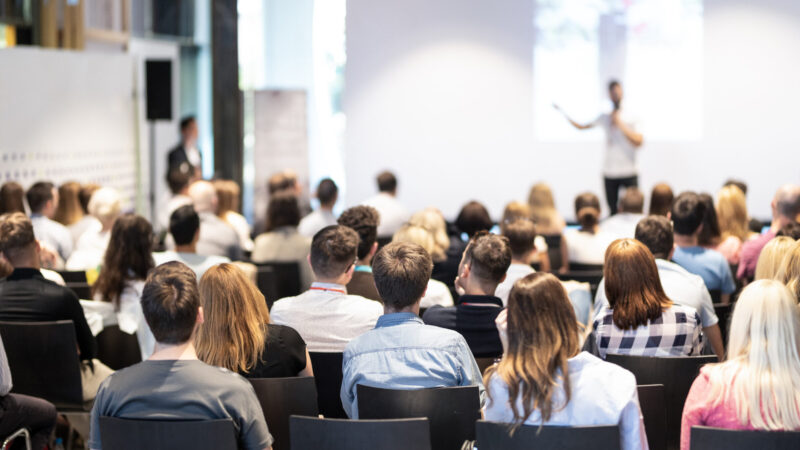 Business speaker giving a talk at business conference event. Speaker giving a talk in conference hall at business event. Audience at the conference hall. Business and Entrepreneurship concept. Focus on unrecognizable people in audience.