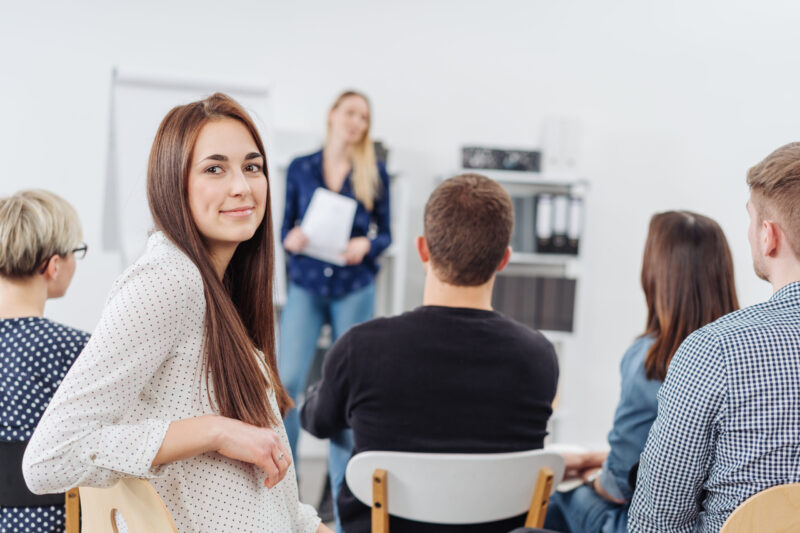 Young woman in a lecture or business presentation turning to smile at the camera viewed from the back of the room