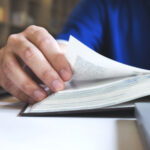 Ein Mann liest in einem Buch  Der Fokus ist auf seiner Hand, mit der er eine Seite umblättert 