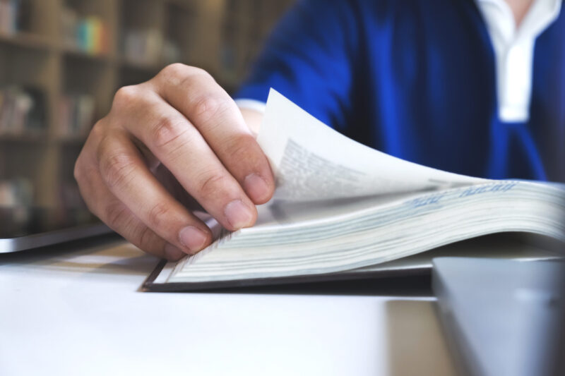 Ein Mann liest in einem Buch. Der Fokus ist auf seiner Hand, mit der er eine Seite umblättert.